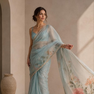 May include: A light blue sheer saree featuring a delicate floral print with soft pink and orange blossoms. The fabric has a subtle shimmer and is finished with a thin, ornate gold border along the edges, paired with a matching sleeveless blouse.