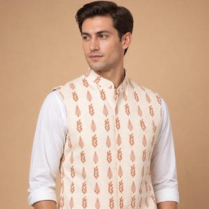 May include: Cream-colored vest with a repeating pattern of reddish-brown leaf-like shapes. The vest has a mandarin collar and a single pocket. The vest is worn over a white long-sleeved shirt and black pants.