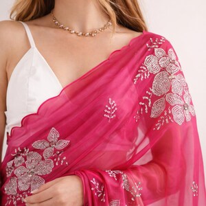 May include: A vibrant fuchsia saree with intricate silver floral embroidery. The sheer fabric drapes over a white top, creating a layered effect. A delicate gold necklace adds a touch of elegance to this traditional garment.