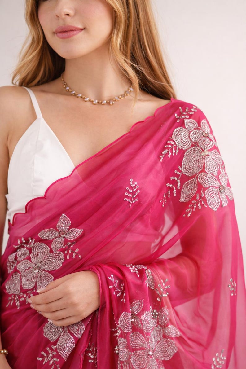 May include: A vibrant fuchsia saree with intricate silver floral embroidery. The sheer fabric drapes over a white top, creating a layered effect. A delicate gold necklace adds a touch of elegance to this traditional garment.