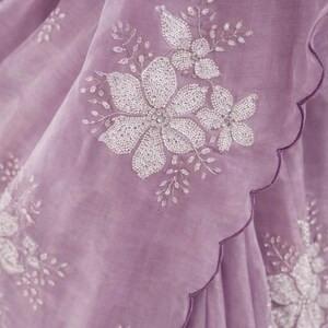 May include: Close-up of a lavender fabric with white floral embroidery. The embroidery features large flowers and smaller floral accents, with a scalloped edge. The fabric appears to be lightweight and sheer.