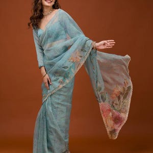 May include: A sheer, light blue organza saree featuring delicate hand-painted floral motifs in soft pink, orange, and green hues. The fabric has a subtle shimmer, and the edges are finished with a thin, metallic gold scalloped border trim.