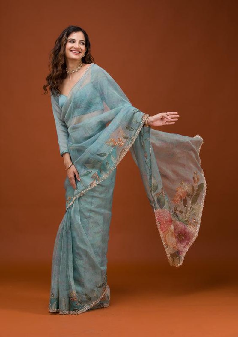 May include: A sheer, light blue organza saree featuring delicate hand-painted floral motifs in soft pink, orange, and green hues. The fabric has a subtle shimmer, and the edges are finished with a thin, metallic gold scalloped border trim.