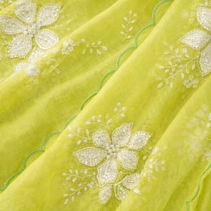 May include: Close-up of a lime green fabric with white floral embroidery. The fabric has a delicate, sheer quality, with a scalloped green edge. The floral design includes detailed petals and leaves, creating a textured appearance.