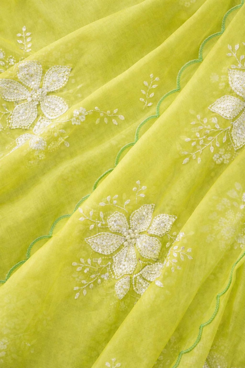 May include: Close-up of a lime green fabric with white floral embroidery. The fabric has a delicate, sheer quality, with a scalloped green edge. The floral design includes detailed petals and leaves, creating a textured appearance.
