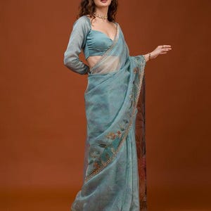 May include: A light blue saree featuring a sheer, translucent fabric with a delicate floral pattern and a decorative border. The garment is paired with a matching long-sleeved blouse that has a sweetheart neckline and a fitted silhouette.