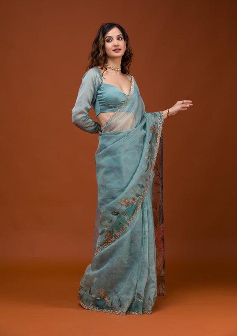 May include: A light blue saree featuring a sheer, translucent fabric with a delicate floral pattern and a decorative border. The garment is paired with a matching long-sleeved blouse that has a sweetheart neckline and a fitted silhouette.