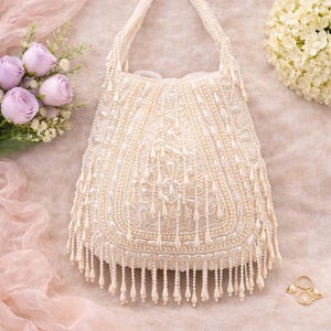 May include: Ivory beaded handbag with a handle. The bag is adorned with intricate beadwork and dangling fringe. The background includes flowers and a pink fabric.