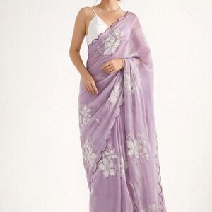 May include: A lavender saree with intricate floral embroidery. The sheer fabric drapes elegantly, featuring a scalloped edge. The saree is paired with a white top. This traditional garment is suitable for special occasions.