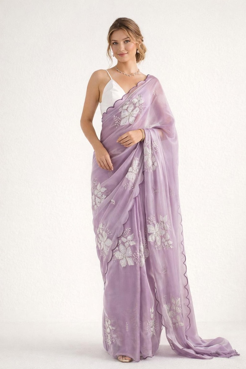 May include: A lavender saree with intricate floral embroidery. The sheer fabric drapes elegantly, featuring a scalloped edge. The saree is paired with a white top. This traditional garment is suitable for special occasions.