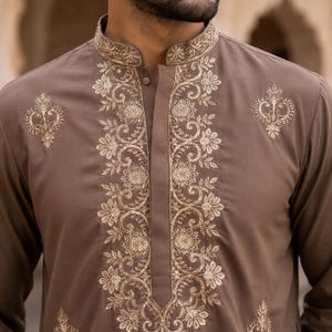 K&ouml;nnte beinhalten: Ein brauner, lang&auml;rmeliger Kurta mit Stehkragen und einer zentralen Knopfleiste. Das Kleidungsst&uuml;ck ist mit aufwendigen, goldfarbenen Blumenstickereien entlang des Ausschnitts, der vorderen Knopfleiste und in symmetrischen Mustern auf der Brust verziert.