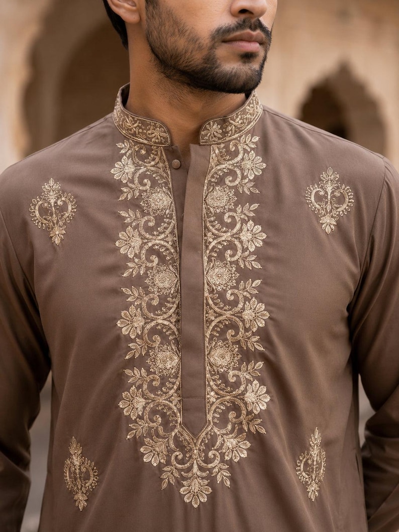 K&ouml;nnte beinhalten: Ein brauner, lang&auml;rmeliger Kurta mit Stehkragen und einer zentralen Knopfleiste. Das Kleidungsst&uuml;ck ist mit aufwendigen, goldfarbenen Blumenstickereien entlang des Ausschnitts, der vorderen Knopfleiste und in symmetrischen Mustern auf der Brust verziert.