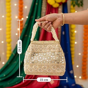 K&ouml;nnte beinhalten: Eine beige Perlenhandtasche, 23 cm breit und 29,5 cm hoch, mit einem perlenartigen Griff. Die Tasche ist mit kleinen Spiegeln und floralen Mustern verziert. Der Hintergrund zeigt farbenfrohe Stoffe und dekorative Lichter.