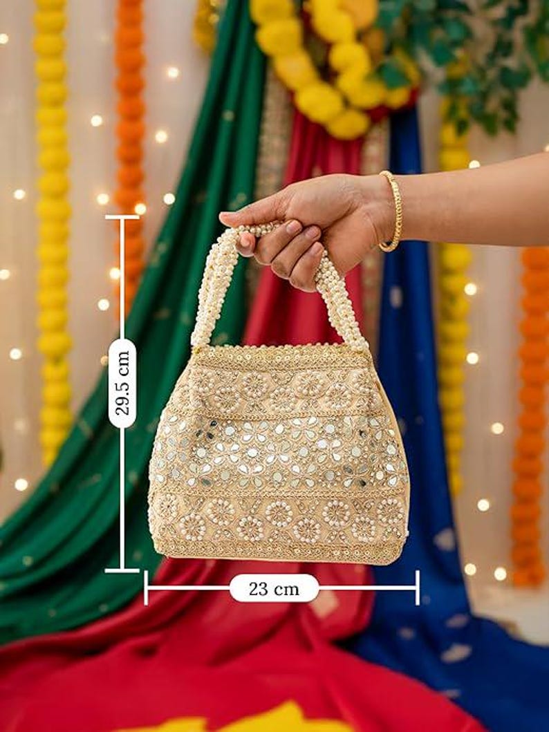 K&ouml;nnte beinhalten: Eine beige Perlenhandtasche, 23 cm breit und 29,5 cm hoch, mit einem perlenartigen Griff. Die Tasche ist mit kleinen Spiegeln und floralen Mustern verziert. Der Hintergrund zeigt farbenfrohe Stoffe und dekorative Lichter.