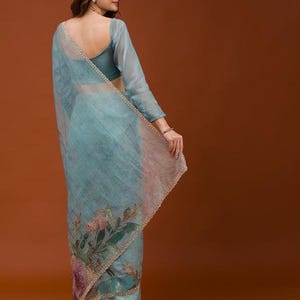 May include: A light blue sheer saree featuring a floral print with pink and green botanical motifs. The fabric has a subtle texture and is finished with a delicate beaded border along the edges. The garment is paired with a matching blue blouse.