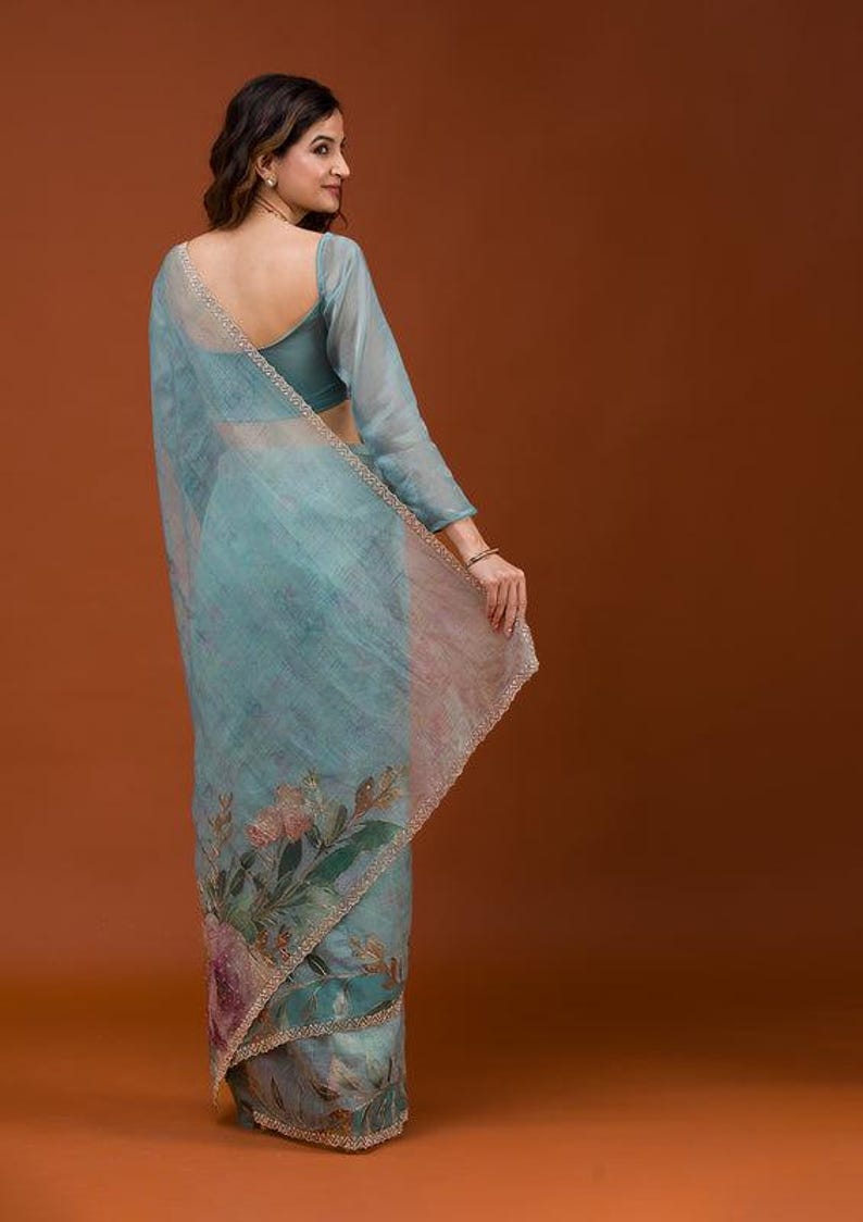 May include: A light blue sheer saree featuring a floral print with pink and green botanical motifs. The fabric has a subtle texture and is finished with a delicate beaded border along the edges. The garment is paired with a matching blue blouse.