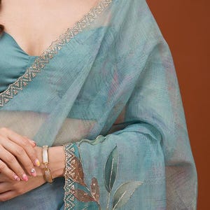 May include: A light blue sheer saree featuring a subtle textured pattern, a decorative border with intricate gold embroidery, and a detailed leaf motif. The garment is paired with a matching solid light blue blouse and a gold-toned choker necklace.