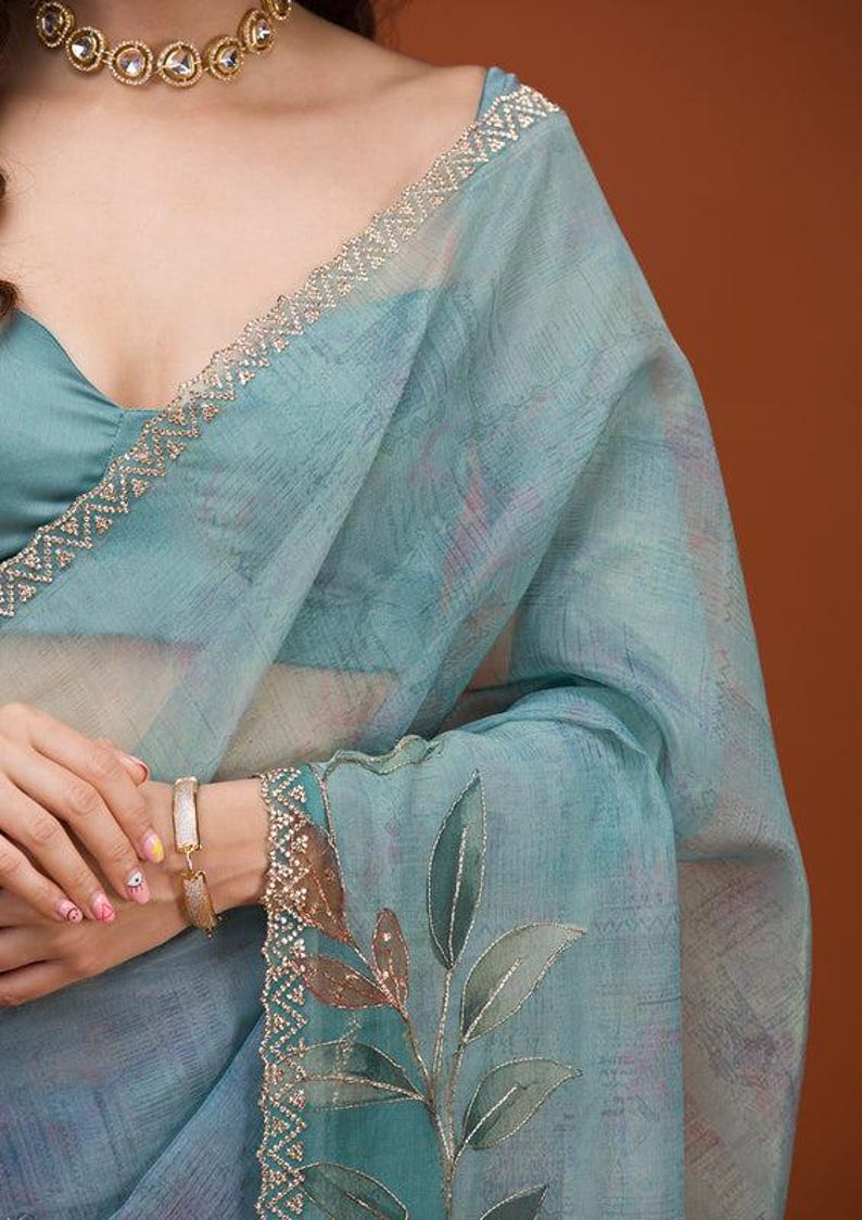 May include: A light blue sheer saree featuring a subtle textured pattern, a decorative border with intricate gold embroidery, and a detailed leaf motif. The garment is paired with a matching solid light blue blouse and a gold-toned choker necklace.