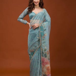 May include: A light blue sheer saree featuring a long-sleeved blouse and a draped skirt with a floral print. The fabric displays a botanical pattern with green leaves and large orange and pink flowers along the border, finished with a thin gold trim.