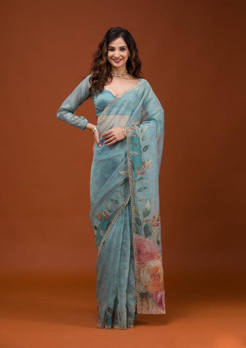 May include: A light blue sheer saree featuring a long-sleeved blouse and a draped skirt with a floral print. The fabric displays a botanical pattern with green leaves and large orange and pink flowers along the border, finished with a thin gold trim.