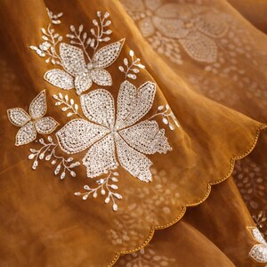 May include: Close-up of a sheer, golden-brown fabric with intricate white floral embroidery. The design features large, detailed flowers and delicate sprigs, with a scalloped edge.
