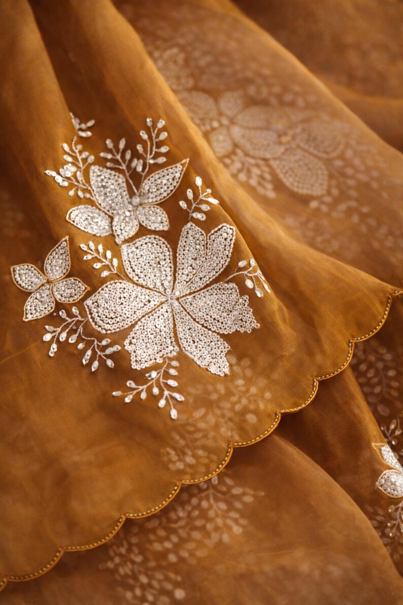 May include: Close-up of a sheer, golden-brown fabric with intricate white floral embroidery. The design features large, detailed flowers and delicate sprigs, with a scalloped edge.
