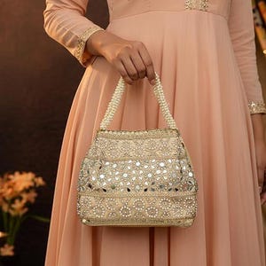 K&ouml;nnte beinhalten: Eine beige verzierte Handtasche mit Perlenhenkel wird von einer Person in einem pfirsichfarbenen Kleid gehalten. Die Tasche weist aufwendige Stickereien und kleine, reflektierende Spiegelakzente auf, wodurch ein dekoratives und elegantes Accessoire entsteht.