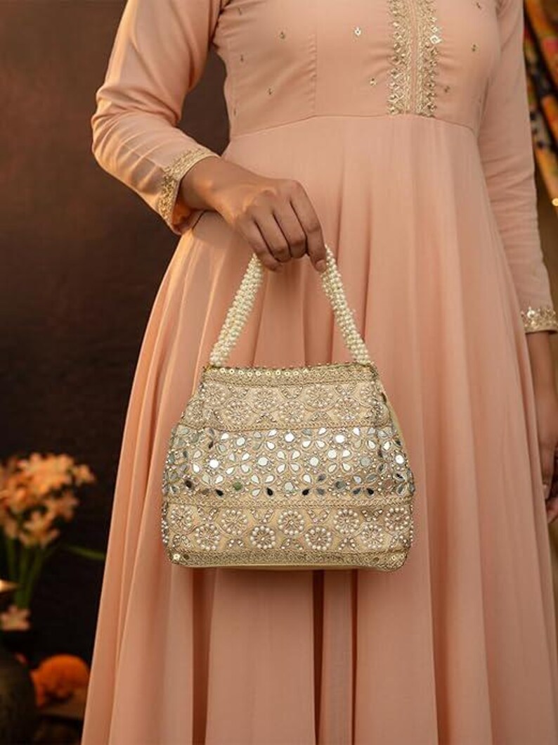 K&ouml;nnte beinhalten: Eine beige verzierte Handtasche mit Perlenhenkel wird von einer Person in einem pfirsichfarbenen Kleid gehalten. Die Tasche weist aufwendige Stickereien und kleine, reflektierende Spiegelakzente auf, wodurch ein dekoratives und elegantes Accessoire entsteht.