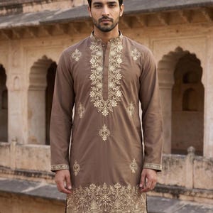 K&ouml;nnte beinhalten: Ein lang&auml;rmeliger brauner Kurta mit aufwendiger goldener Stickerei entlang des Stehkragens, der Knopfleiste und des Saums. Das Kleidungsst&uuml;ck hat eine gerade Silhouette mit einem gekn&ouml;pften Ausschnitt und passenden goldenen Blumenmustern.