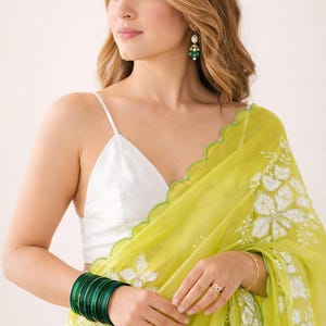 May include: A woman wearing a white spaghetti strap top and a light green sari with white floral embroidery. She is wearing green bangles, earrings, and a ring. The sari has a scalloped edge.
