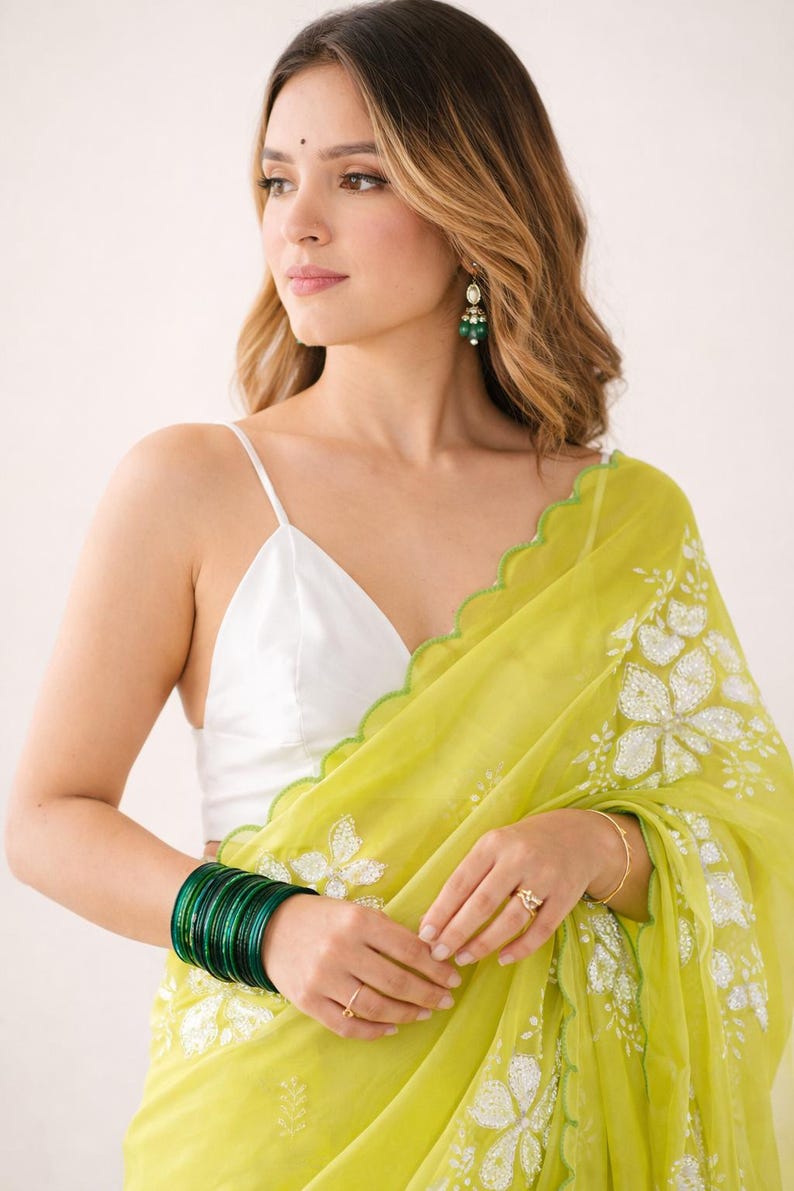 May include: A woman wearing a white spaghetti strap top and a light green sari with white floral embroidery. She is wearing green bangles, earrings, and a ring. The sari has a scalloped edge.