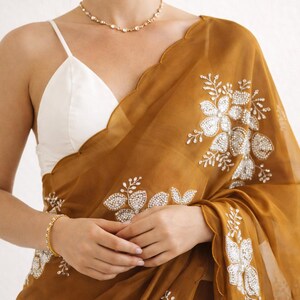 May include: Mustard-colored sari with intricate floral embroidery. The sheer fabric drapes over a white camisole. A gold bracelet and necklace add subtle sparkle. The sari features a scalloped edge.