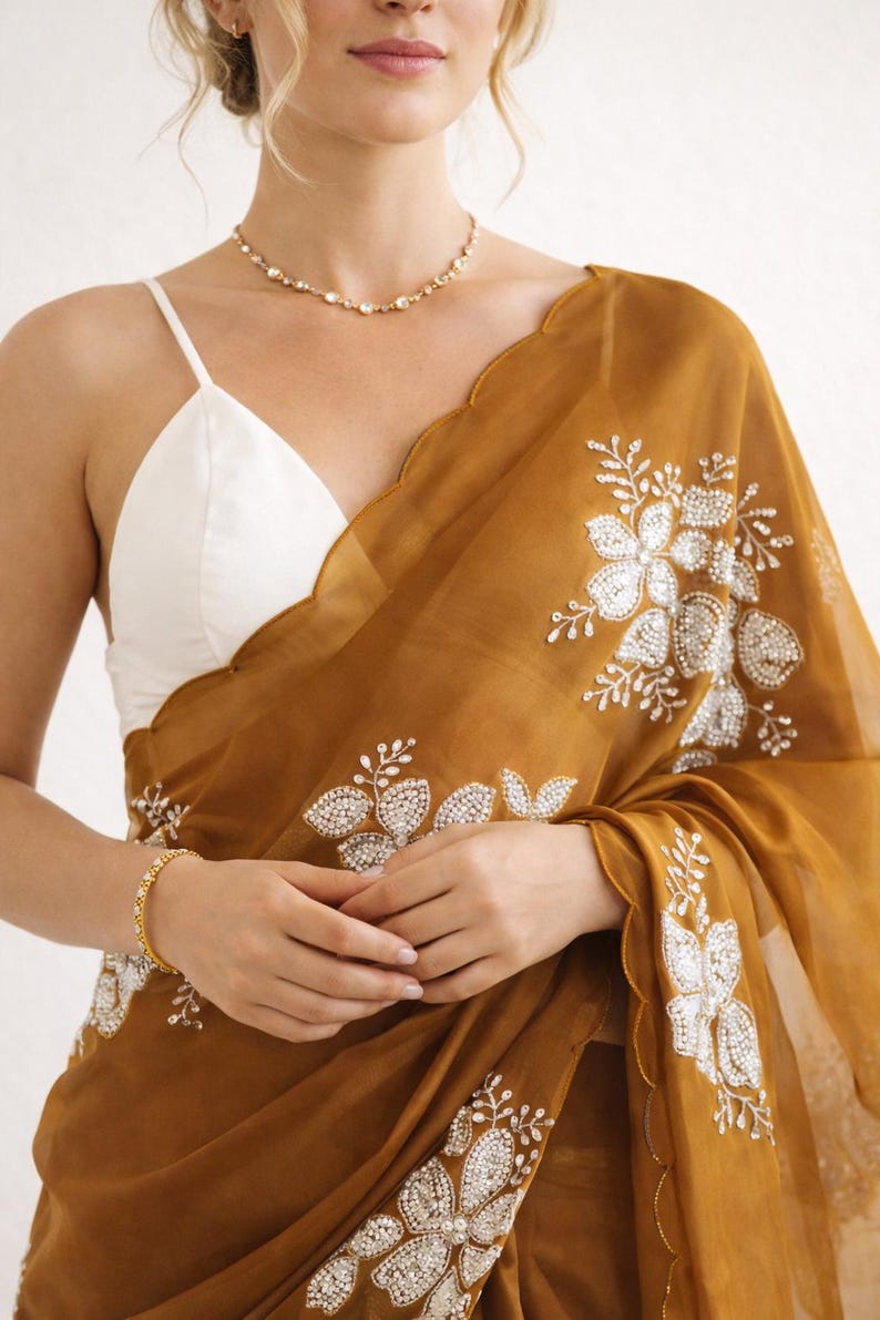 May include: Mustard-colored sari with intricate floral embroidery. The sheer fabric drapes over a white camisole. A gold bracelet and necklace add subtle sparkle. The sari features a scalloped edge.