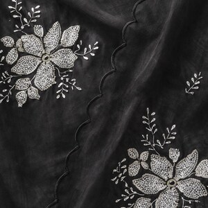 May include: Black fabric with silver floral embroidery. The design features large, detailed flowers and smaller leaf accents, all embellished with sparkling silver beads. A scalloped edge adds a decorative touch to the fabric.