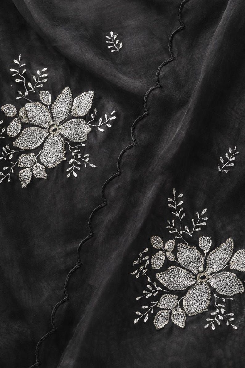 May include: Black fabric with silver floral embroidery. The design features large, detailed flowers and smaller leaf accents, all embellished with sparkling silver beads. A scalloped edge adds a decorative touch to the fabric.