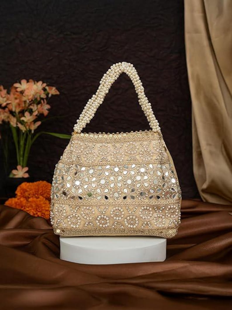 K&ouml;nnte beinhalten: Beige Handtasche mit Perlenhenkel und aufwendigen Verzierungen. Die Tasche hat ein Blumenmuster mit kleinen Spiegeln und perlenartigen Perlen. Der Griff besteht aus kleinen, wei&szlig;en Perlen. Die Tasche wird auf einem wei&szlig;en Podest pr&auml;sentiert.