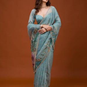 May include: A sheer, light blue organza saree featuring a delicate floral print in shades of green, purple, and brown. The fabric has a subtle shimmer and is finished with a decorative, intricate gold-toned border along the edges of the drape.