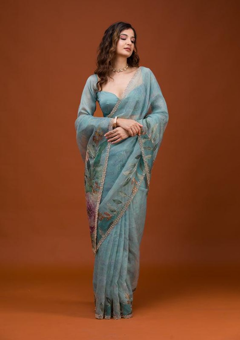 May include: A sheer, light blue organza saree featuring a delicate floral print in shades of green, purple, and brown. The fabric has a subtle shimmer and is finished with a decorative, intricate gold-toned border along the edges of the drape.