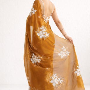 May include: Mustard-colored sari with white floral embroidery. The sheer fabric drapes over the shoulders and features a scalloped edge. A white, open-back top is visible beneath the sari.