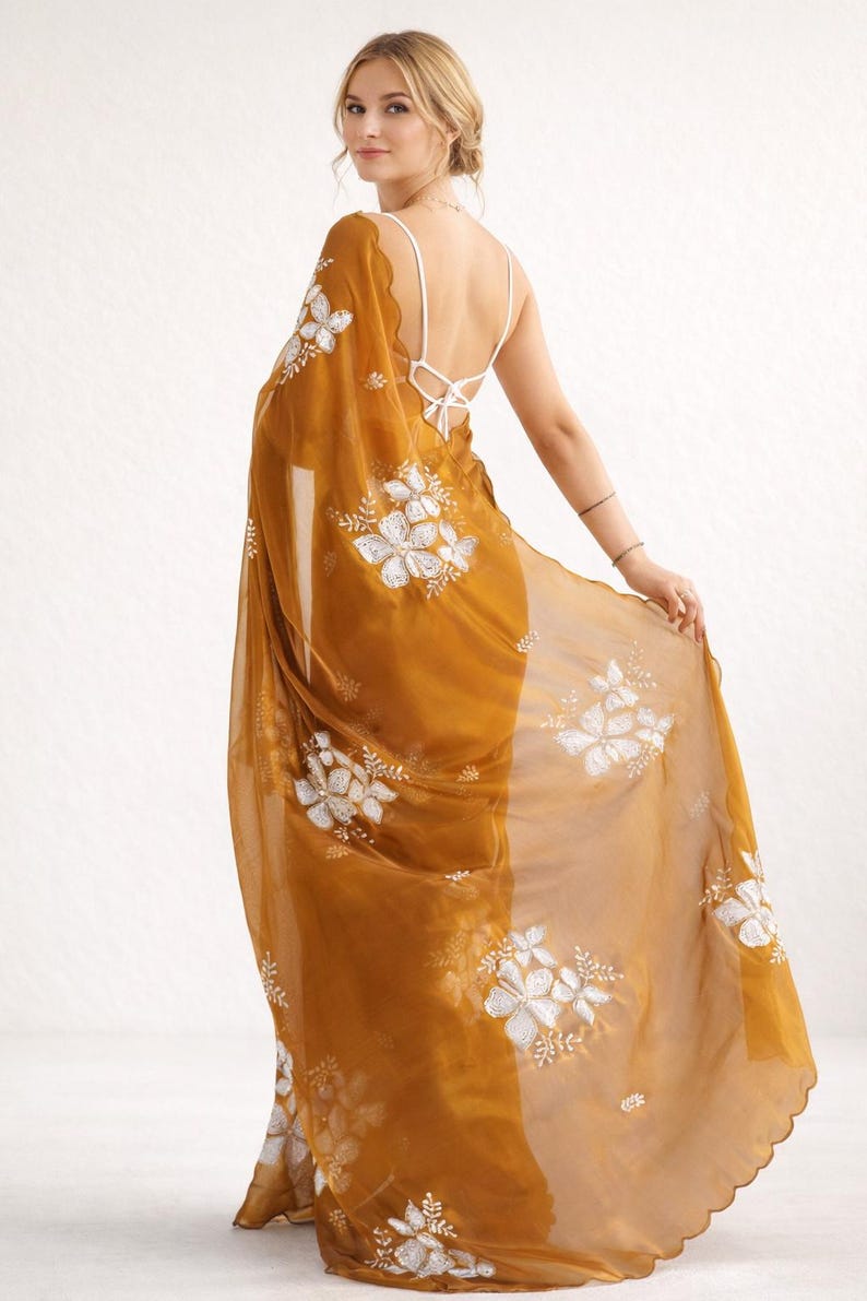May include: Mustard-colored sari with white floral embroidery. The sheer fabric drapes over the shoulders and features a scalloped edge. A white, open-back top is visible beneath the sari.