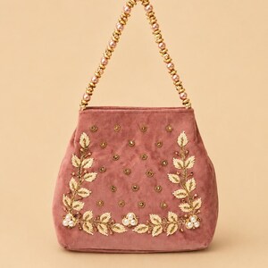 Puede incluir: Bolso de mano de terciopelo rosa empolvado con adornos dorados y perlas. El bolso presenta un asa de cuentas y está adornado con hojas doradas y detalles de perlas. Un accesorio elegante para ocasiones especiales.