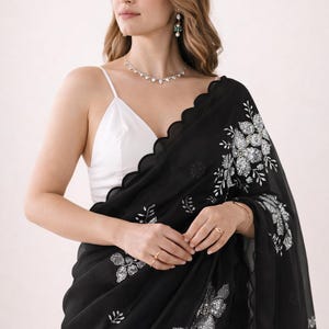 May include: Black saree with silver floral embroidery. The saree has a scalloped edge and is paired with a white camisole. The saree is a traditional garment, often worn for special occasions.
