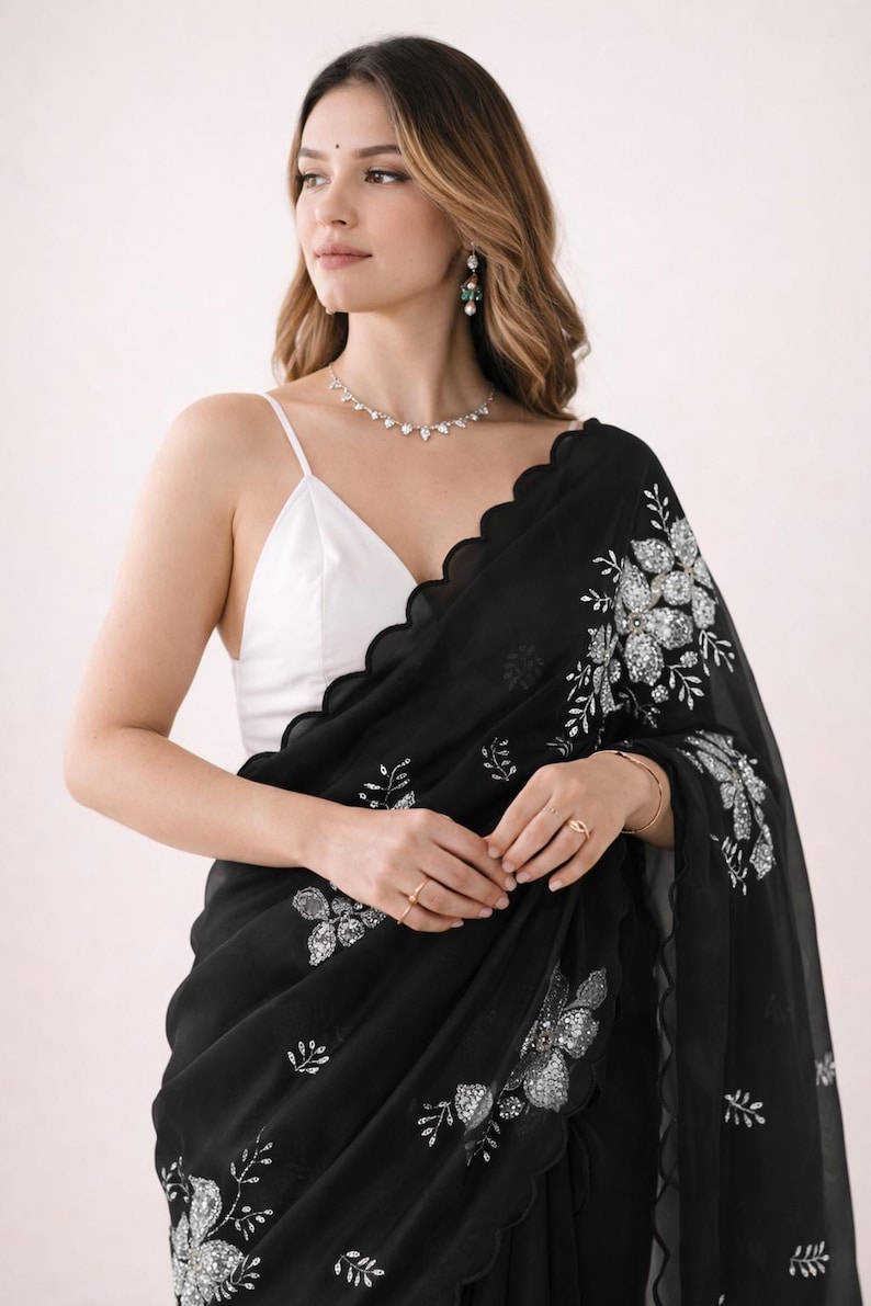 May include: Black saree with silver floral embroidery. The saree has a scalloped edge and is paired with a white camisole. The saree is a traditional garment, often worn for special occasions.