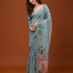 May include: A light blue sheer saree featuring a floral print with large pink and orange flowers along the border. The garment has long sleeves, a V-neckline, and a decorative gold-toned trim along the edges of the fabric and the blouse.