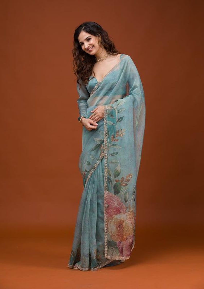 May include: A light blue sheer saree featuring a floral print with large pink and orange flowers along the border. The garment has long sleeves, a V-neckline, and a decorative gold-toned trim along the edges of the fabric and the blouse.
