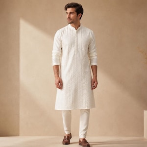 Puede incluir: Una kurta de color blanco roto con mangas largas, cuello mao y rayas verticales texturizadas. La prenda se combina con pantalones a juego en el mismo tono y mocasines de cuero marrón, creando un conjunto coordinado de estética neutra.