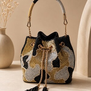 May include: A bucket bag covered in black, gold, and silver beads arranged in a camouflage pattern. The bag features a drawstring closure with gold-toned hardware and two long beaded tassels. It has a single handle with black and silver beads.