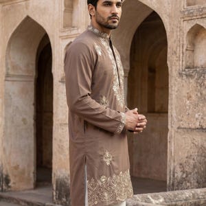 K&ouml;nnte beinhalten: Ein lang&auml;rmeliger brauner Kurta mit aufwendiger cremefarbener Stickerei entlang des Stehkragens, der Knopfleiste, der &Auml;rmelb&uuml;ndchen und des unteren Saums, kombiniert mit einer schlichten wei&szlig;en Hose.