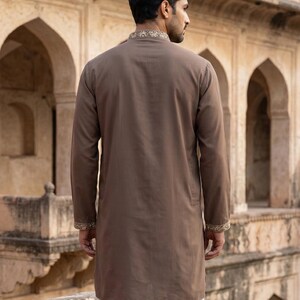 K&ouml;nnte beinhalten: Ein lang&auml;rmeliger, einfarbiger brauner Kurta mit Stehkragen und Manschetten, die mit aufwendigen, hellen Stickereien verziert sind. Das Kleidungsst&uuml;ck wird &uuml;ber einer schlichten wei&szlig;en Hose getragen und zeigt eine klare, strukturierte Silhouette.