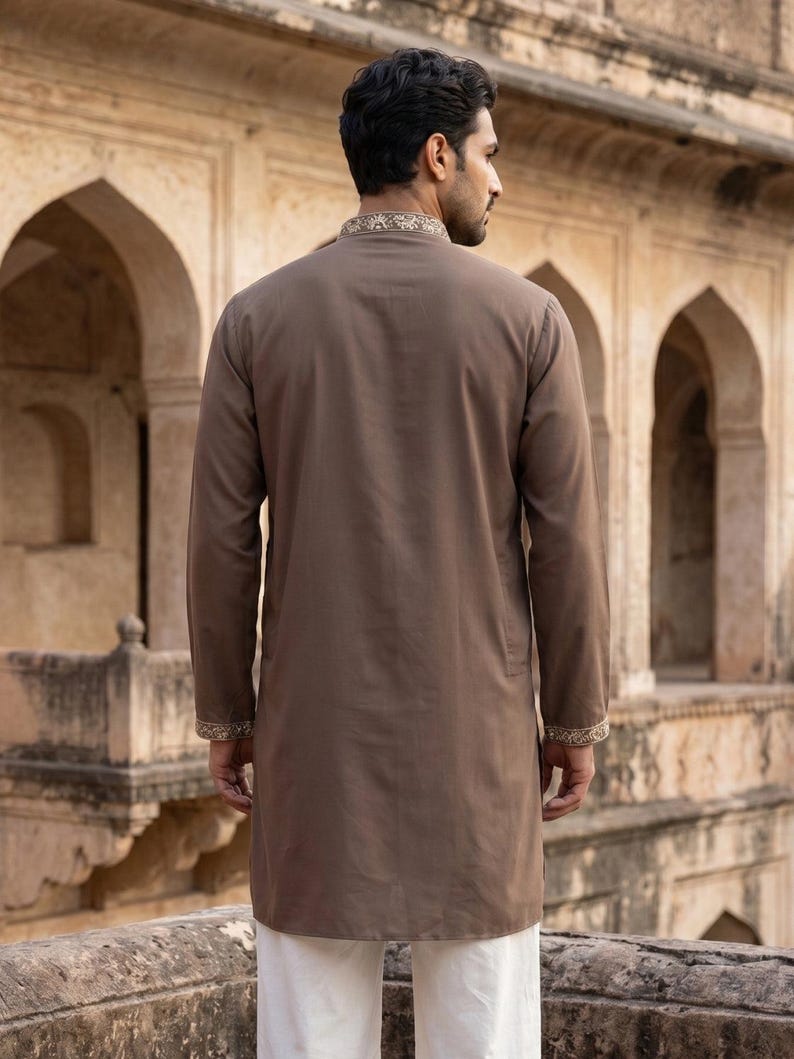 K&ouml;nnte beinhalten: Ein lang&auml;rmeliger, einfarbiger brauner Kurta mit Stehkragen und Manschetten, die mit aufwendigen, hellen Stickereien verziert sind. Das Kleidungsst&uuml;ck wird &uuml;ber einer schlichten wei&szlig;en Hose getragen und zeigt eine klare, strukturierte Silhouette.